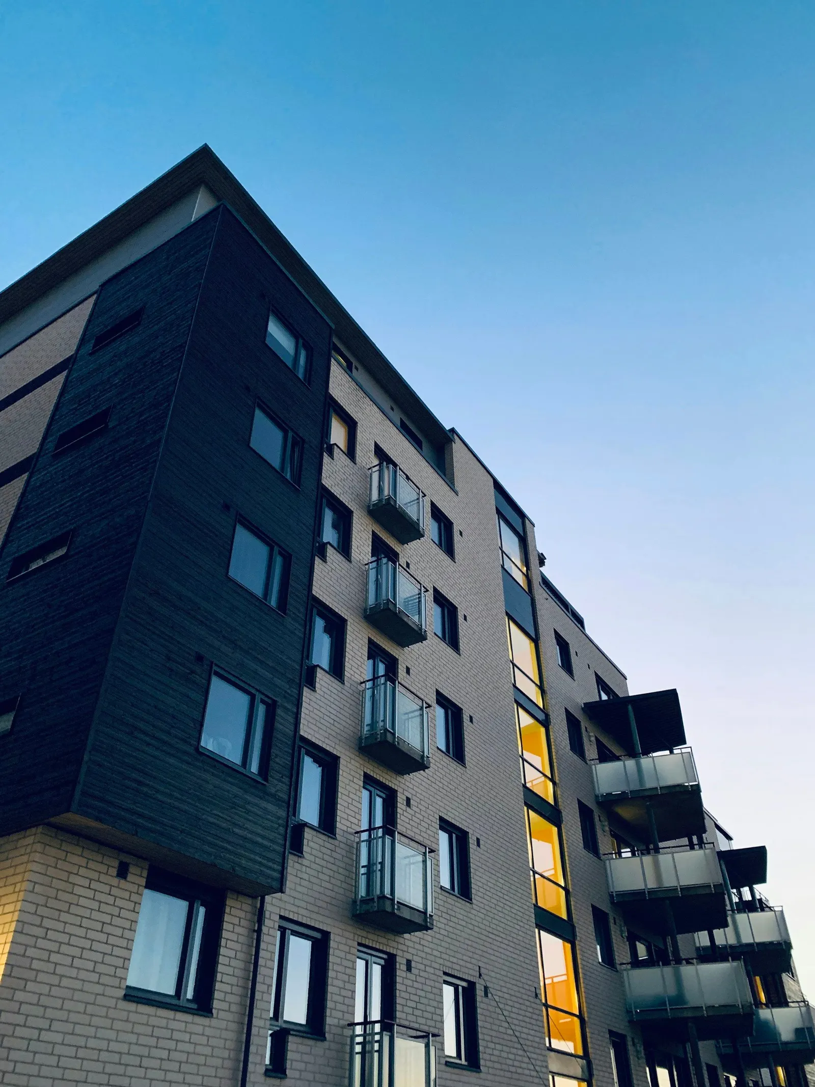 Moderne boligblokk med balkonger og mørk trekledning, sett nedenfra mot blå himmel